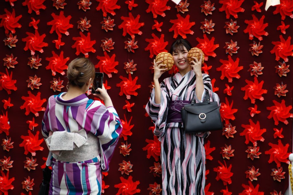 Une femme en kimono, souriante, tient des pâtisseries devant un fond floral coloré.