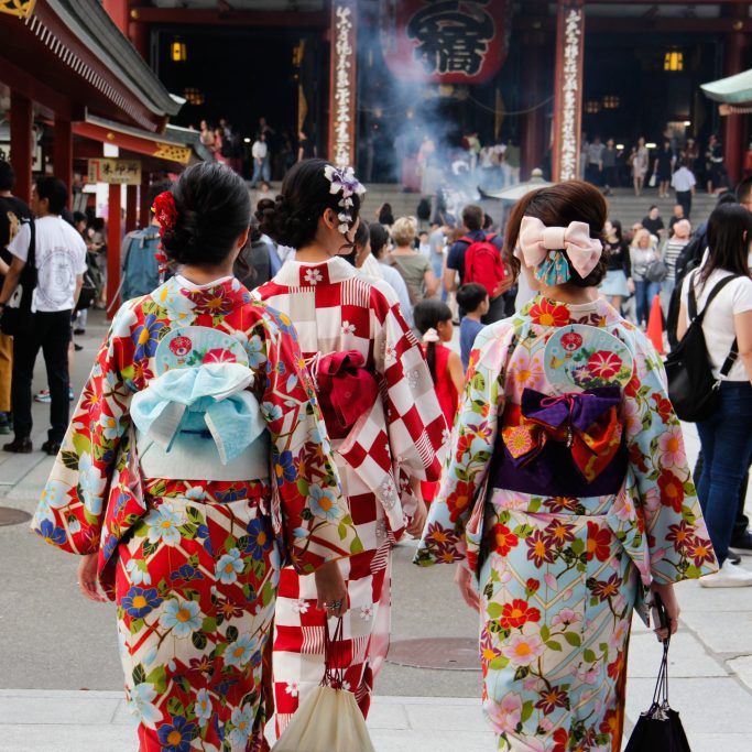Trois femmes en kimonos colorés marchent vers un temple japonais, entourées de visiteurs.