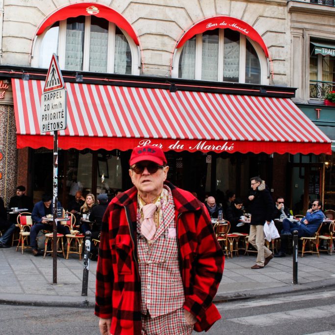 Homme en manteau à carreaux rouges devant un café animé avec auvent rayé.